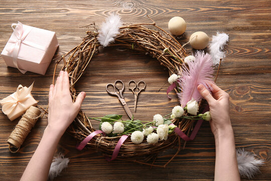 Woman making Easter wreath with eggs, feathers and flowers on brown wooden background