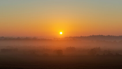 Naklejka premium Golden sunrise over misty rural landscape with soft haze and distant trees