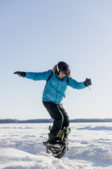 Person riding electric unicycle monowheel on frozen snow-covered lake field in winter landscape with blue sky forest background wearing safety helmet goggles, extreme outdoor adventure sport activity © MarijaBazarova