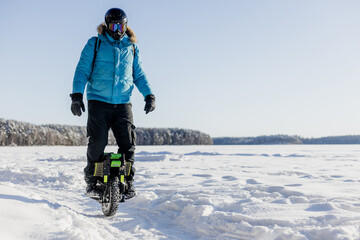 Person riding electric unicycle monowheel on frozen snow-covered lake field in winter landscape with blue sky forest background wearing safety helmet goggles, extreme outdoor adventure sport activity © MarijaBazarova