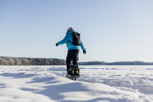 Person riding electric unicycle monowheel on frozen snow-covered lake field in winter landscape with blue sky forest background, extreme outdoor adventure sport activity