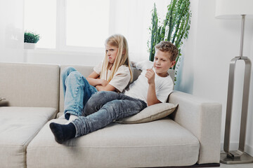 Obraz premium Upset girl and relaxed boy sitting apart on couch in bright living room. Concept of sibling conflict, family tension, and childhood emotions indoors.