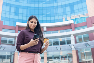 Fototapeta premium Young indian businesswoman checking her smart phone and holding a disposable coffee cup standing outside a modern corporate office building. Corporate job. Copy space. Technology.