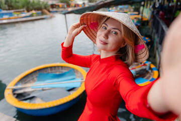 Fototapeta premium Young caucasian female tourist in red ao dai making selfie photo by coconut basket boats in hoi an, vietnam.