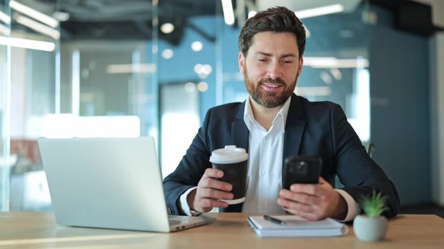Professional man smiles, happily interacting with smartphone while enjoying coffee in modern office. Positive emotions expressed. Connection, communication, success, and joyful work atmosphere.