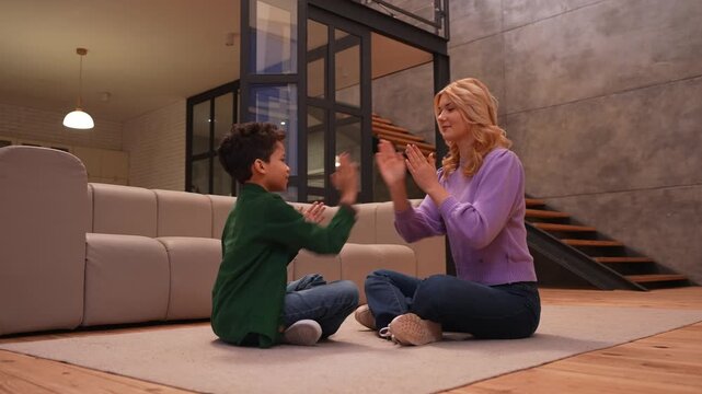 Cheerful lovely teenage nanny and excited cute African American school age boy sitting cross legged on floor , having fun playing clapping game while enjoying leisure in domestic room.