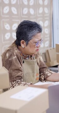 Senior asian businesswoman frowns, reviews accounting paperwork, checks online order on laptop, compares invoice with receipt, notes tracking code, worries shipping delay, studio workday drags.