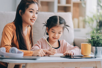 Obraz premium A cheerful Asian mother helps her young daughter do homework and color a book at home, while the father works on a laptop in the background. A cozy, bright, and modern family lifestyle.