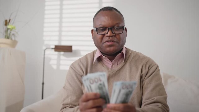 Happy middle-aged african american man counting new hundred dollars bills. Medium portrait of black male person in home interior, salary and fee for good job, successful financial deal of businessman