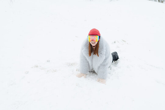 Playful winter scene of a woman in a grey fur coat and rainbow ski goggles crawling on snow for winter sports advertising, banner and background