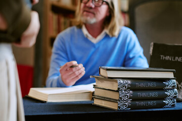 Middle aged Caucasian man sitting at table signing books during presentation event, stack of...