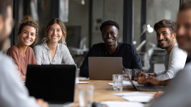 A group of people are smiling and sitting around a table with laptops. They seem to be having a good time and enjoying each other's company