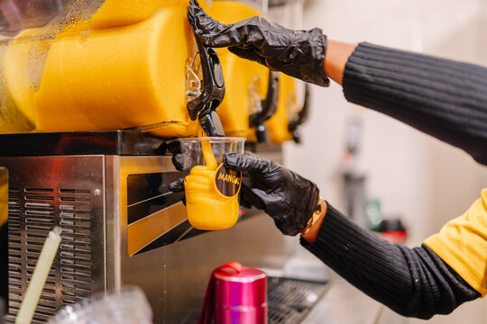 Person in black gloves pours mango smoothie from a slush machine into a clear cup at a beverage stand