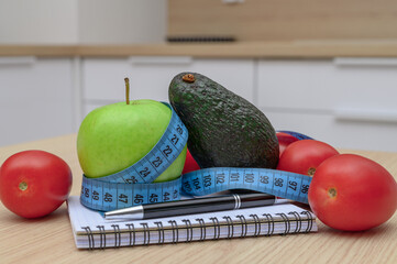 Ripe tomatoes lying on a table next to an avocado and a green apple wrapped in a measuring tape