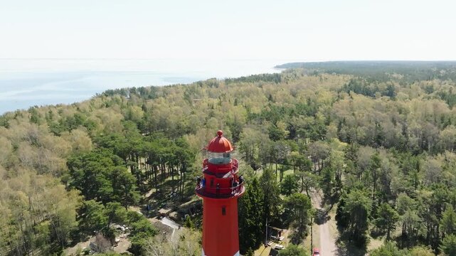 Aerial drone view of a red and white lighthouse on the Baltic Sea coast. Smooth flyover above the tower surrounded by forest with sea horizon on a sunny day.
