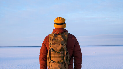 Man wearing yellow beanie and backpack stands with back to camera, gazing across a vast frozen snowy landscape under clear morning sky, peaceful winter exploration © Dmitrii
