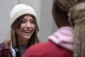 Female friends chatting in narrow corridor with ribbed wall, wearing cream beanie and headphones