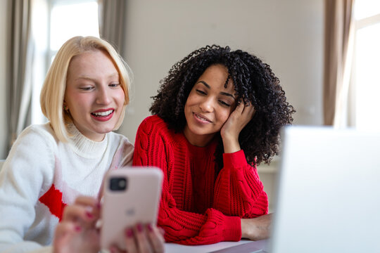Two Young Women Looking at Smartphone and Smiling at Home. Diverse Friends Using Mobile App and Laptop Concept. Happy Female Roommates Browsing Social Media on Cellphone in Modern Living Room.
