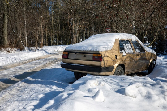 An old gold sedan sits abandoned and heavily covered in thick winter snow in a wooded park.