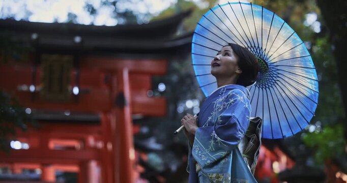 Smiling, beautiful Japanese woman in kimono holding umbrella as she stands near torii gates - slow motion