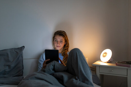 Young woman lying in bed at home at night using tablet