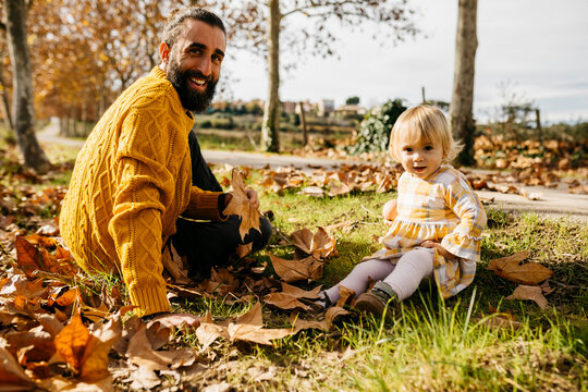 Father and daughter sitting on meadow with autumn leaves, morning day in the park