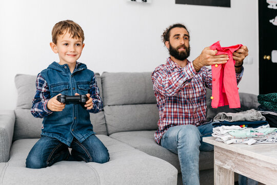 Portrait of smiling little boy playing computer game while his father folding laundry