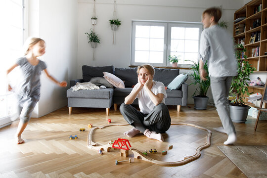 Stressed out mother sitting in the middle of toys, while children are running around her
