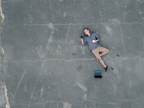 Young businessman with coffee to go lying on tarmac using mini tablet, quadcopter view