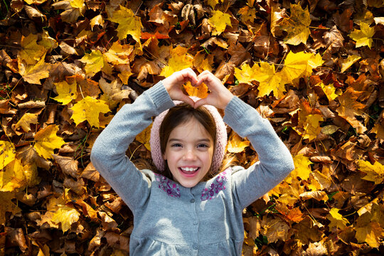 Portrait of happy little girl lying on autumn leaves shaping heart with her hands