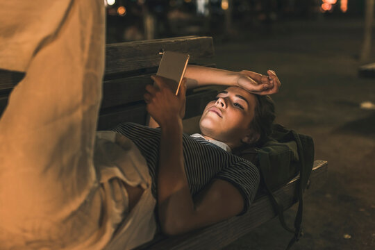 Young woman lying on bench at night using cell phone