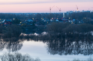 Old countryside houses on the bank of the river and urban modern residential houses construction with cranes on the background, sunrise view, eclectic residential areas background