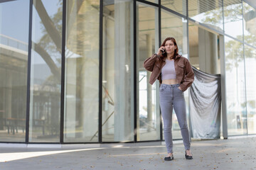 Obraz premium Woman standing outdoors engaging in a phone conversation, using her mobile device for communication and networking within a contemporary architectural business district