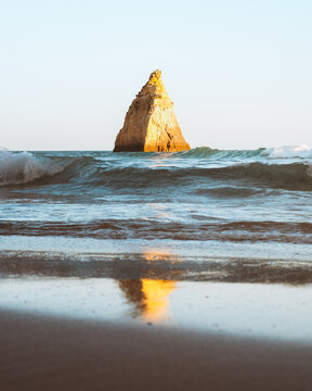 View of a solitary rock formation rises majestically from the turquoise sea, its golden hue mirrored in the wet sand, kissed by gentle waves, Alvor, Faro, Portugal.