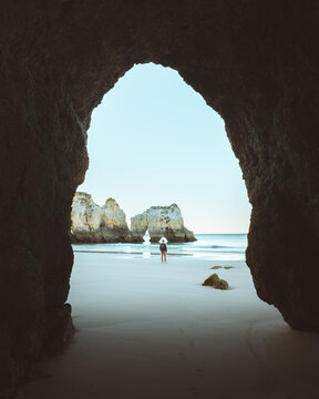 View of the sandy beach beckons through the dark, cool cave opening, framing a lone figure against the azure sky and sea, Alvor, Faro, Portugal.