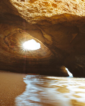 View of sunbeams piercing through the eroded cave opening onto the sandy beach, with gentle waves lapping at the shore, creating a serene coastal scene, Lagoa, Portugal.