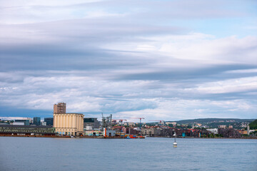 Industrial area of Oslo is seen from across the water, featuring warehouses, cranes, and tall silos. Cloudy sky and city backdrop, Norway mix of urban industry and coastal setting