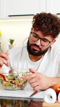 Arabian man forces salad bites in kitchen feeling tired of strict keto diet and healthy nutrition at home. Young guy bored with a diet routine craving comfort food while trying weight loss lifestyles