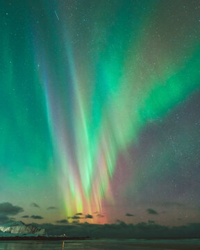 View of the vibrant aurora borealis dancing across the sky, casting ethereal green and purple hues on the shoreline, mirroring in the tranquil waters, Ramberg, Nordland, Norway.