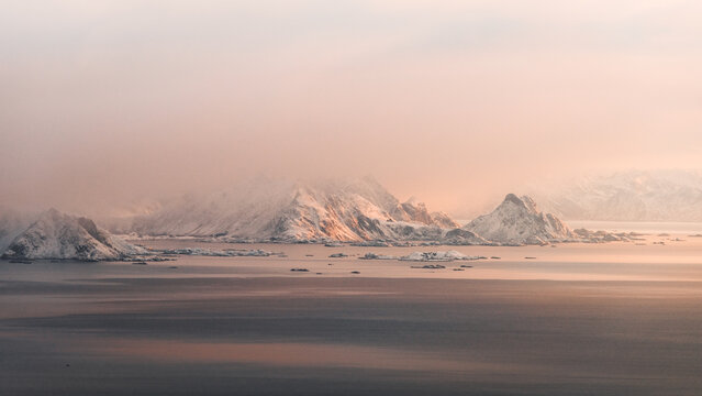 View of snow-capped peaks rise majestically from the tranquil waters, embracing the soft, diffused light of dawn, creating a serene arctic scene, Reine, Nordland, Norway.