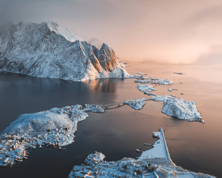 View of snow-dusted islands connected by bridges under a soft, ethereal light, with imposing, snow-capped mountains looming in the background, Reine, Nordland, Norway.
