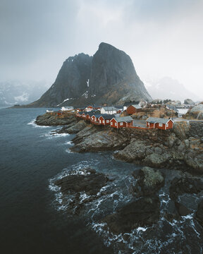 View of crimson cabins nestle against the rugged, snow-dusted peaks, where the moody sea whispers tales of ancient voyages, Hamnoy, Nordland, Norway.
