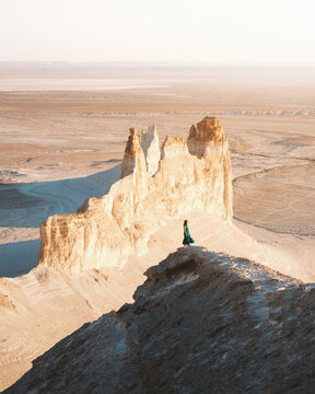 View of a solitary figure in a teal dress atop a windswept ridge, gazing at the stark beauty of sun-baked cliffs and boundless desert plains, Aktau, Mangystau Region, Kazakhstan.