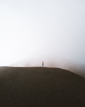 View of a solitary figure standing atop a dark, textured hill, enveloped in a thick, ethereal fog, blending earth and sky in Nicolosi, Sicily, Italy.