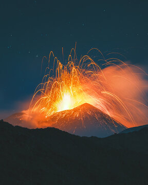 View of fiery sparks erupting from a volcano, painting the night sky with incandescent light against a dark foreground, Nicolosi, Sicily, Italy.