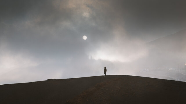 View of a solitary figure stands silhouetted against a hazy sky, the sun a pale orb veiled by clouds in Nicolosi, Sicily, Italy.