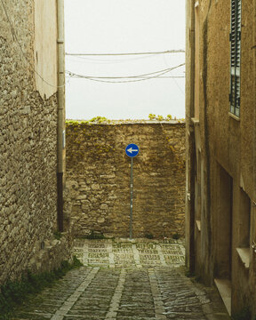 View of a narrow cobblestone street descends between aged stone buildings, guided by a blue directional sign against a weathered wall, Erice, Sicily, Italy.