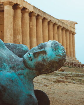 View of the fallen Icarus statue with its striking blue-green patina against the backdrop of ancient Greek temple columns, Agrigento, Sicily, Italy.
