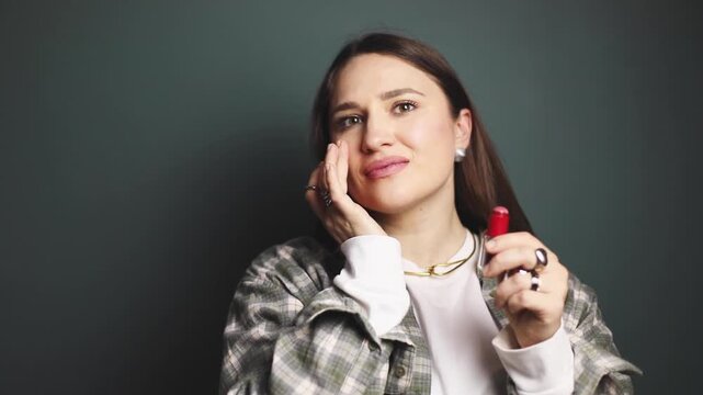 Smiling brunette woman applying bright blush stick on cheekbones and nose, blending with finger, natural makeup routine in motion, beauty cosmetic product demonstration on gray green background studio
