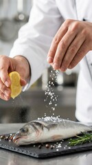 Chef's hand squeezing lemon over fresh fish on a black slate plate, with salt and pepper being sprinkled, garnished with rosemary, in a modern kitchen setting